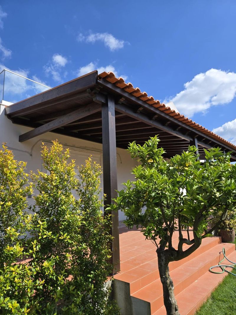 Alpendre com telhado de telha e estrutura de madeira junto a um jardim.