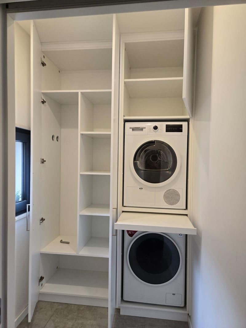 Armário de lavandaria branco aberto, com máquina de lavar e máquina de secar sobrepostas e prateleiras ao lado.